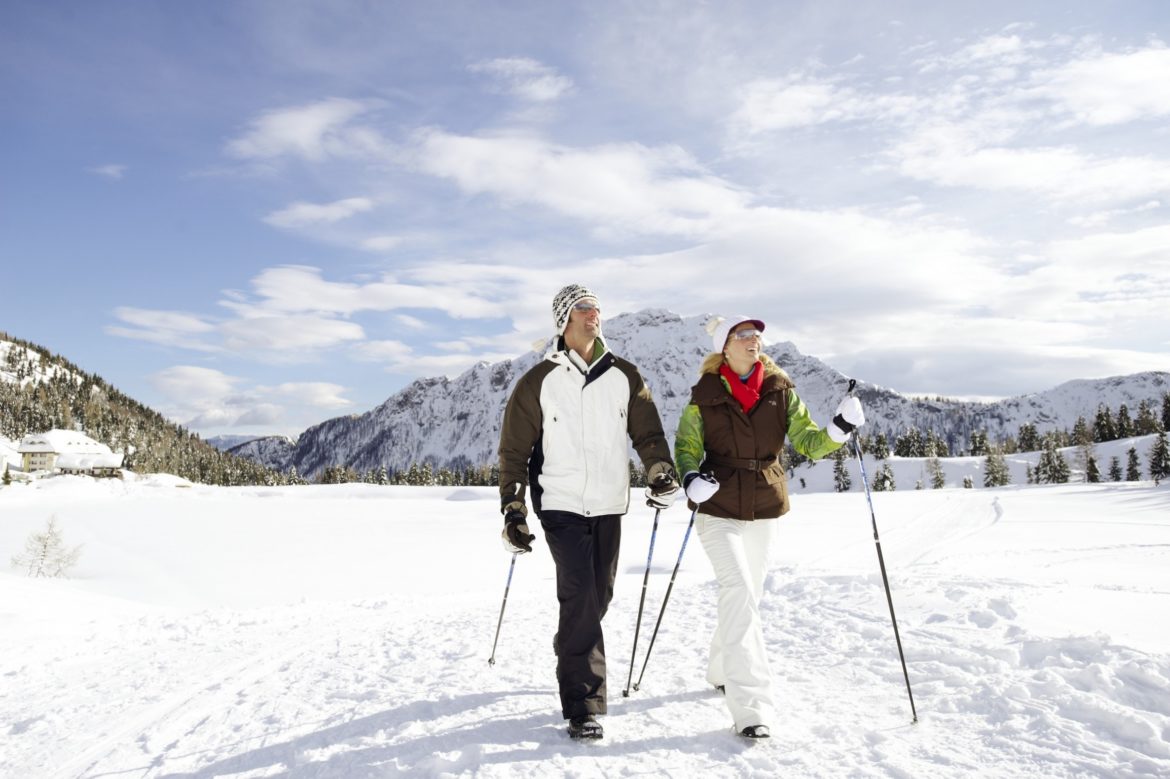 Paar mit Wanderstöcken auf einem verschneiten Weg vor Bergen in der Sonne