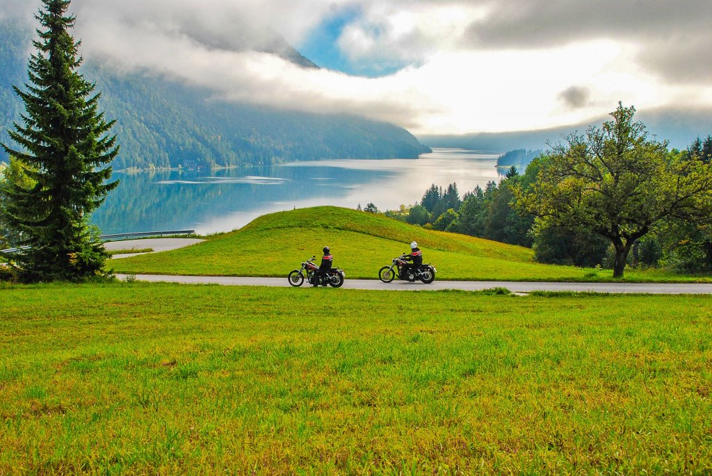 Zwei Motorradfahrer auf einer Straße zwischen Wiese und Seeufer, im Hintergrund Berge