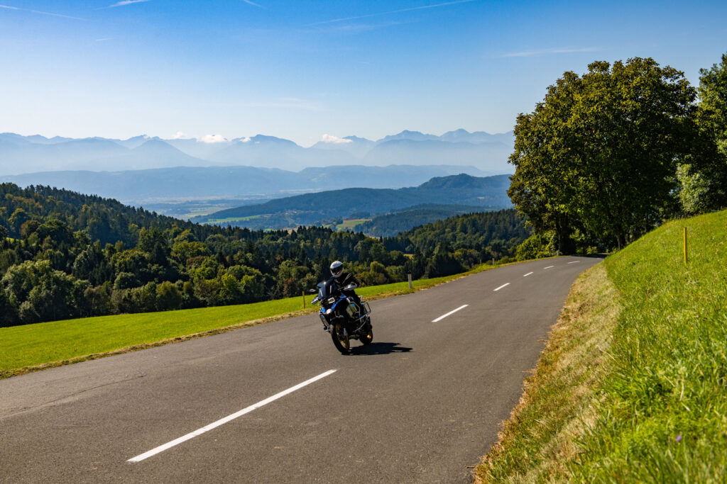 Motorradfahrer auf einer leeren Landstraße am Waldrand mit Bergpanorama