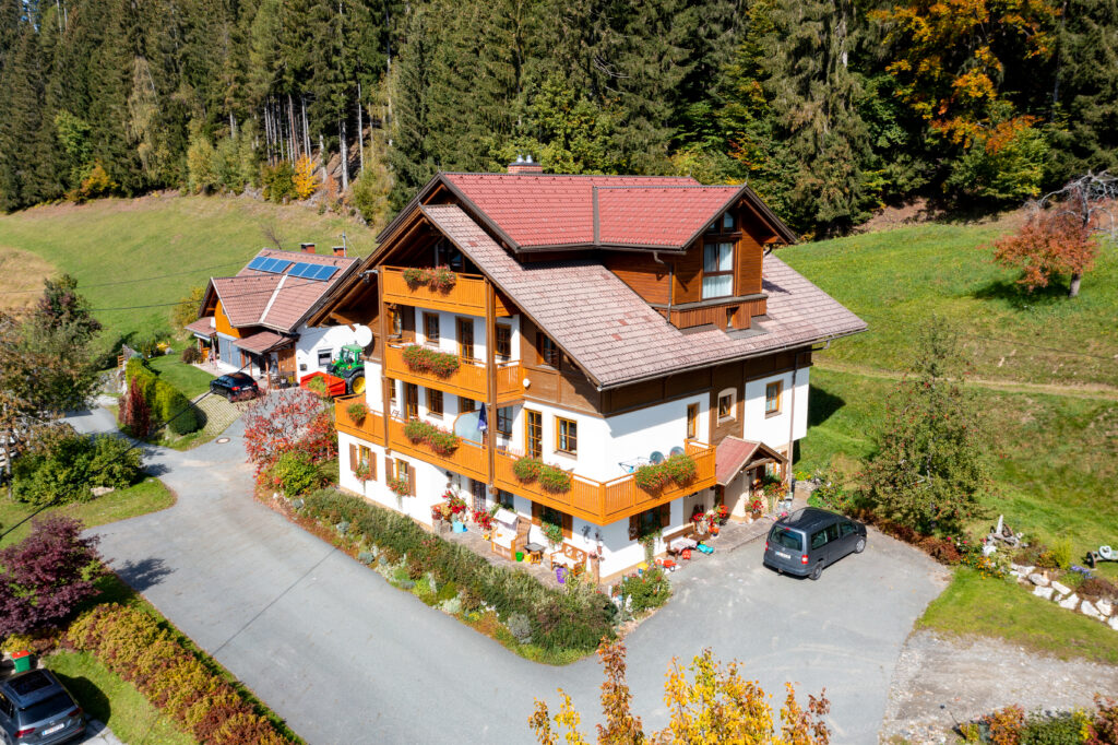 Luftaufnahme eines Hauses mit Holzbalkonen und Blumenkästen beim Berghotel Presslauer, umgeben von Wiesen und Wald