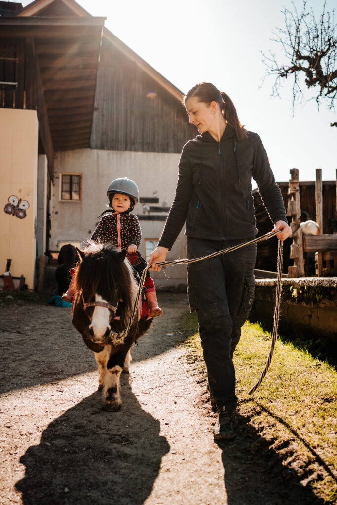 Kind reitet auf einem Pony, eine Frau führt es über den Hof